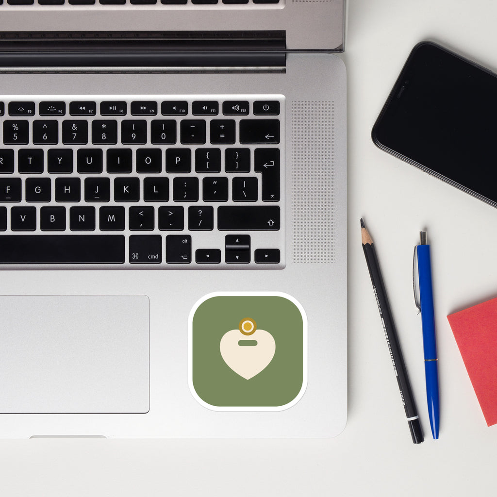 The Amor Budget app icon sticker stuck to a laptop palm rest beside the trackpad, with a phone, pencil, pen and red notepad on a white desk.